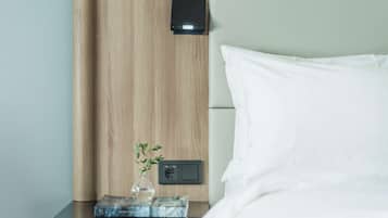 Una cama bien hecha con sábanas blancas, cabecera de madera, mesita de noche con un florero de plantas verdes y un pequeño dispositivo negro en la pared.