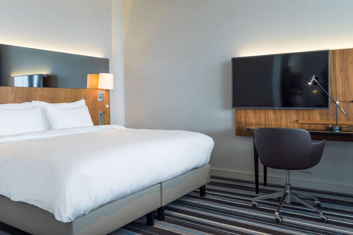 A hotel room with a bed, a desk with a chair, a TV, and a lamp.