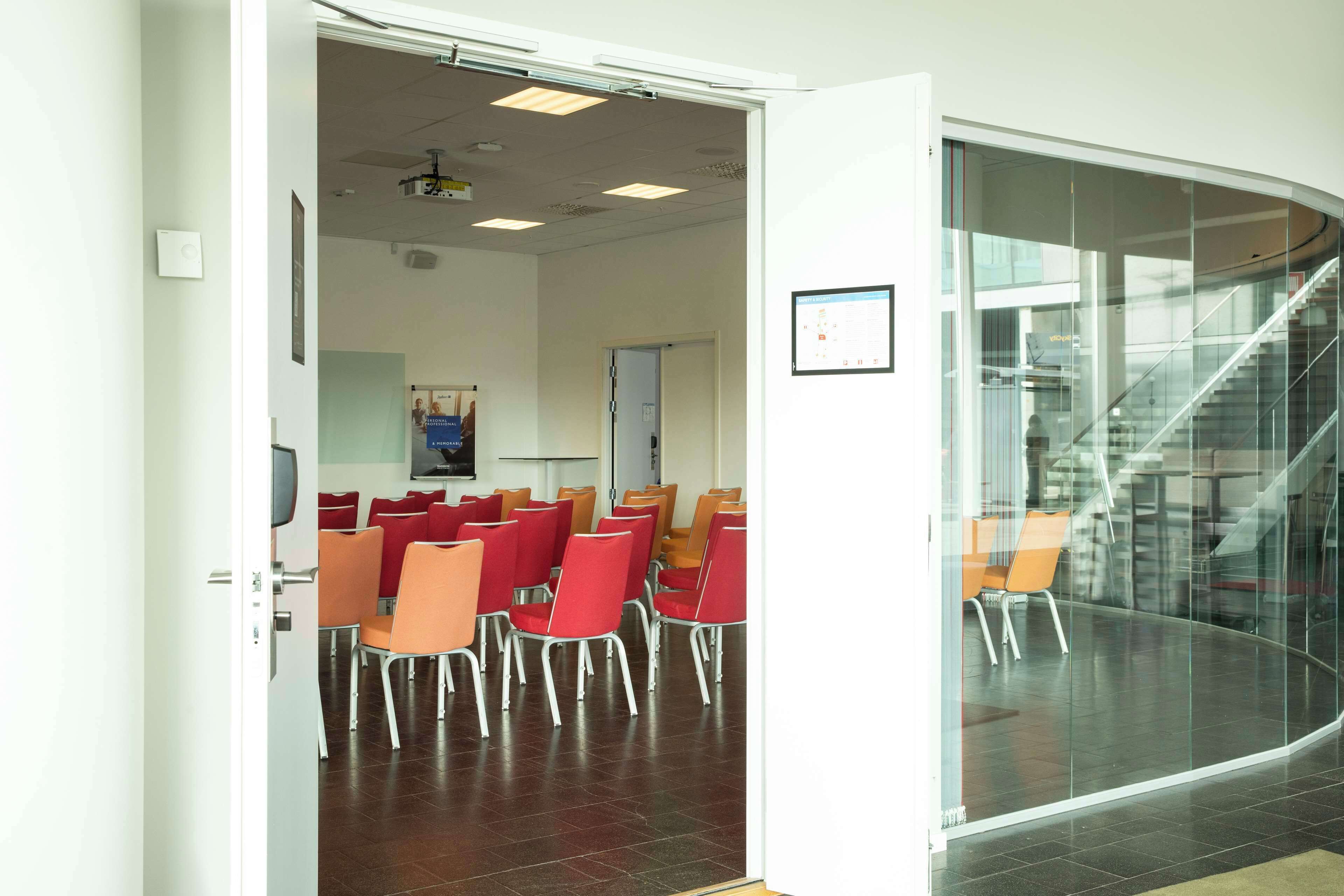 Un espace intérieur comprenant une salle de conférence, des chaises rouges et orange, un tableau blanc et un escalier visible à travers une paroi en verre.