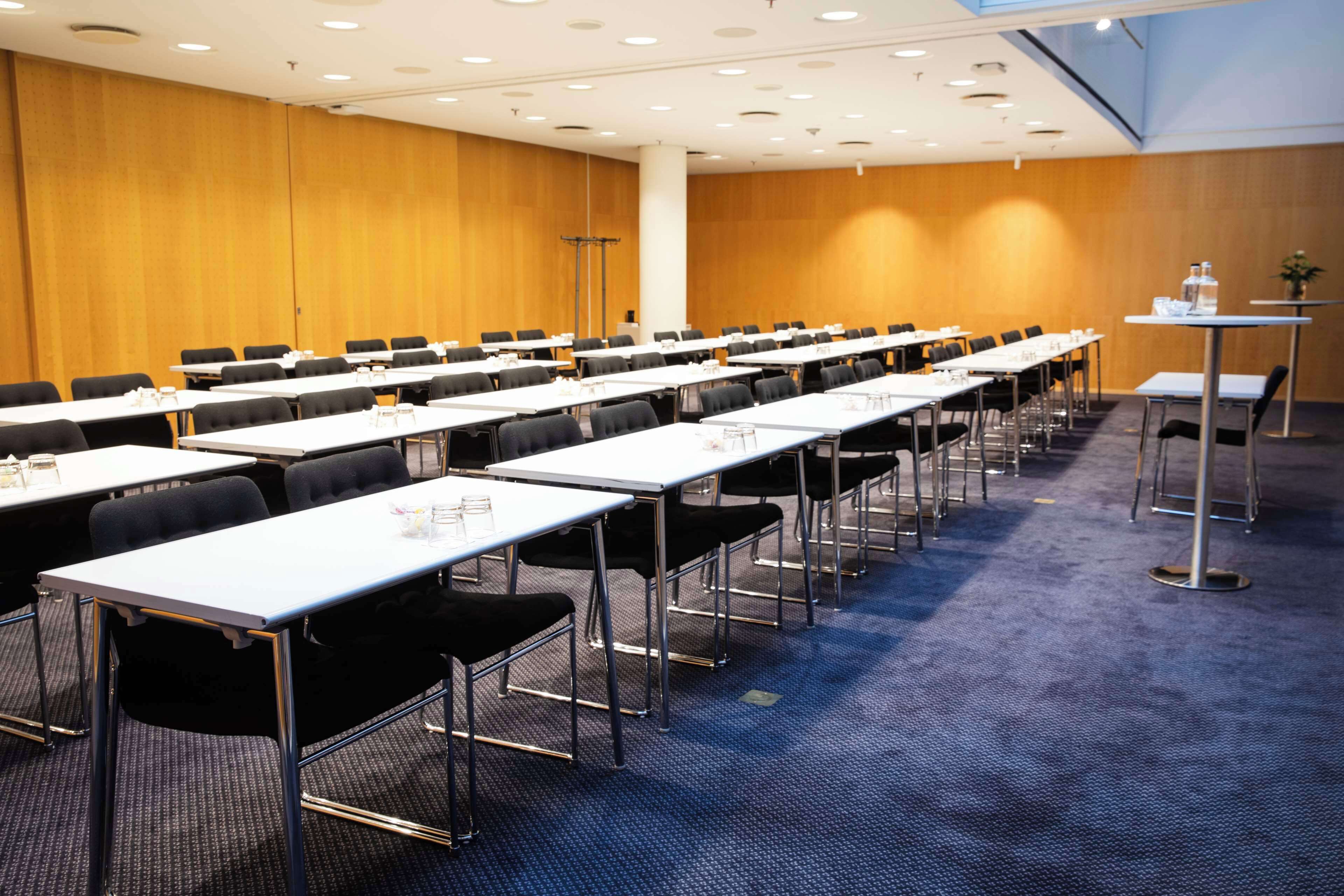 A conference room with rows of tables and chairs, a carpeted floor, and wooden panel walls.