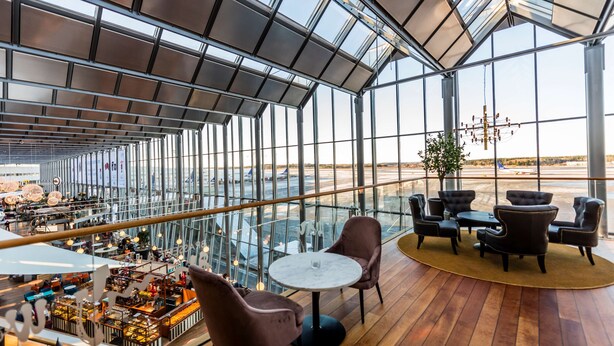 A spacious indoor area with a high ceiling, large windows, and a view of an airport. There are several seating areas with tables and chairs.