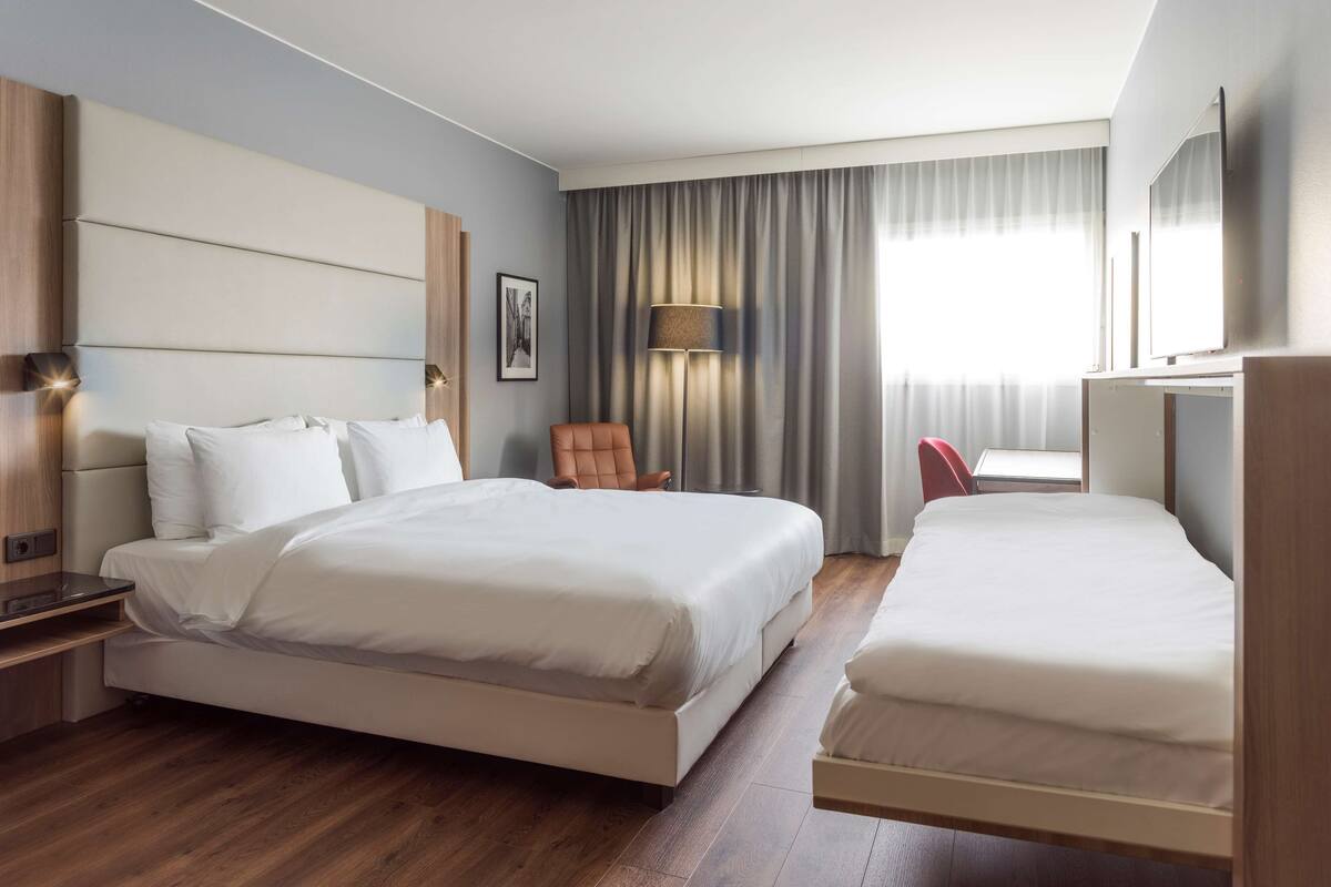 A hotel room with a large bed, a smaller bed, a desk, a chair, a lamp, and a window with curtains.