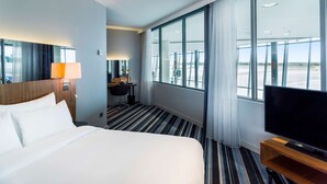 A hotel room with a large bed, a desk with a lamp, a television, and large windows offering a view of the airport.
