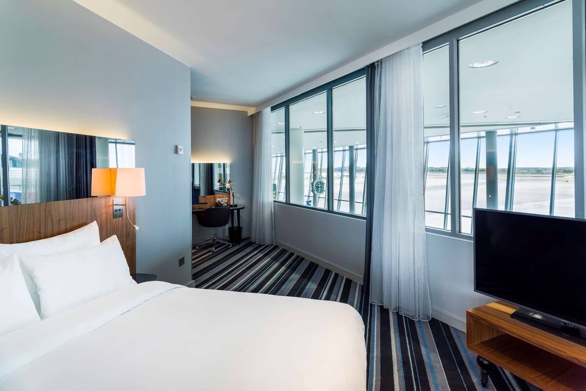 A hotel room with a large bed, a desk with a lamp, a television, and large windows offering a view of the airport.