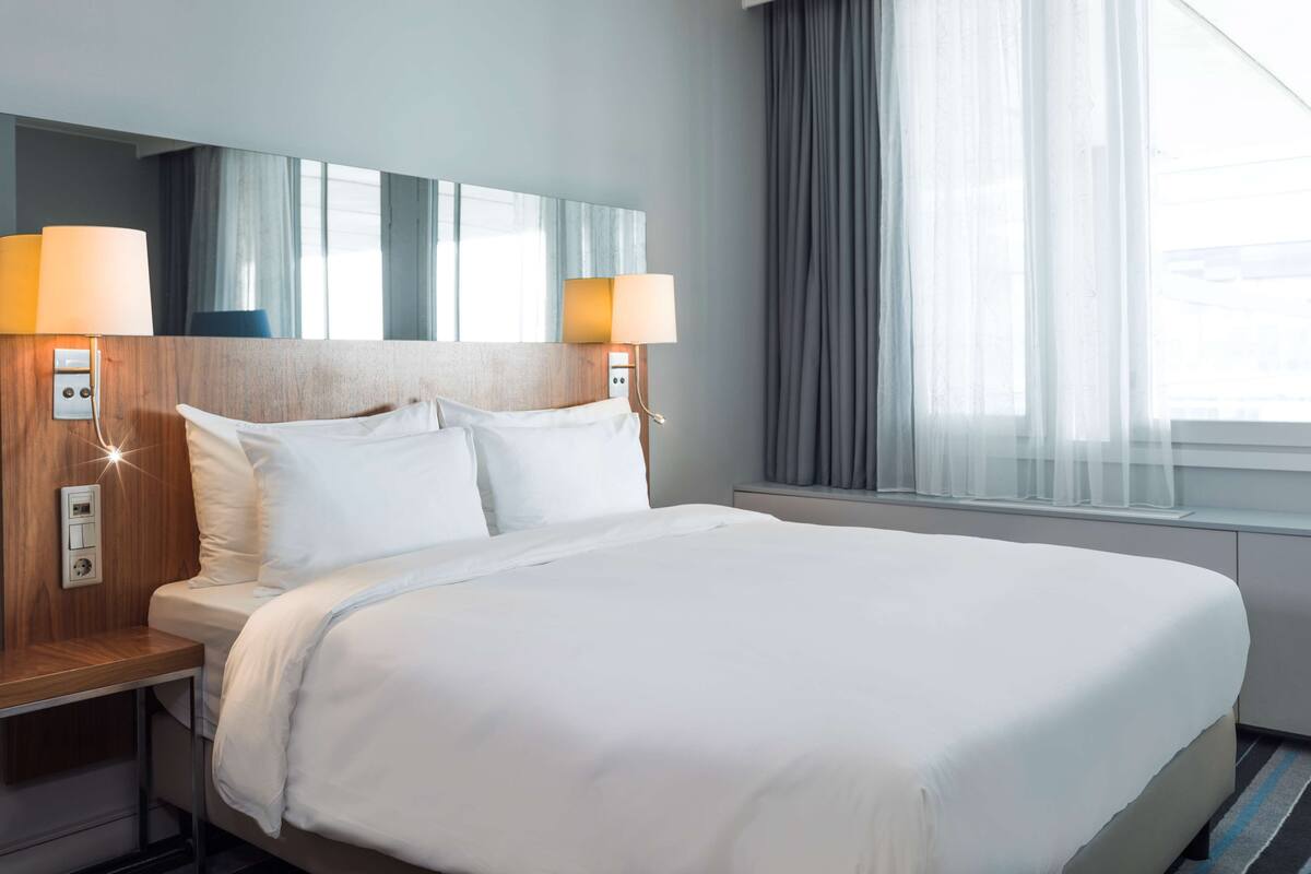 A hotel room with a large bed, two bedside lamps, a nightstand, a window with curtains, and a mirror above the bed.