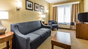 A hotel room with a dark blue sofa, a wooden coffee table, a TV on a wooden stand, a side table with a lamp, and a window with curtains.
