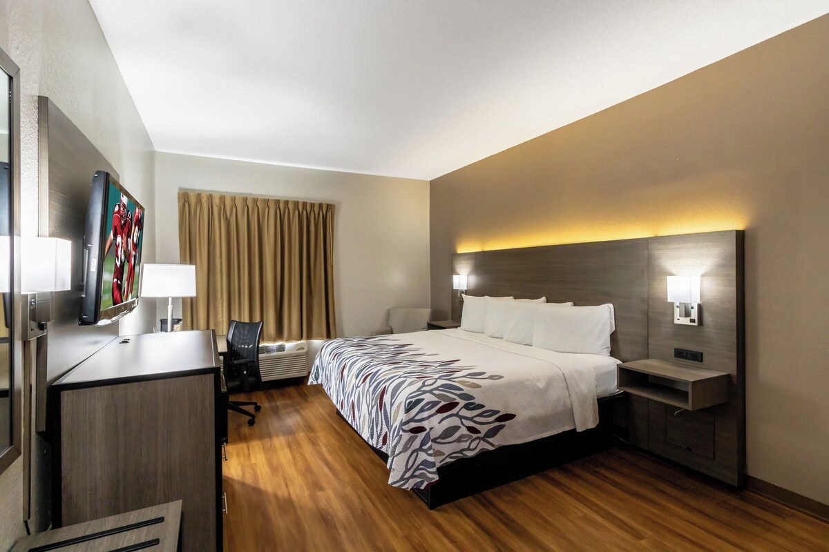 A hotel room with a bed, bedside tables, a desk with a chair, a flat-screen TV, and a window with curtains.