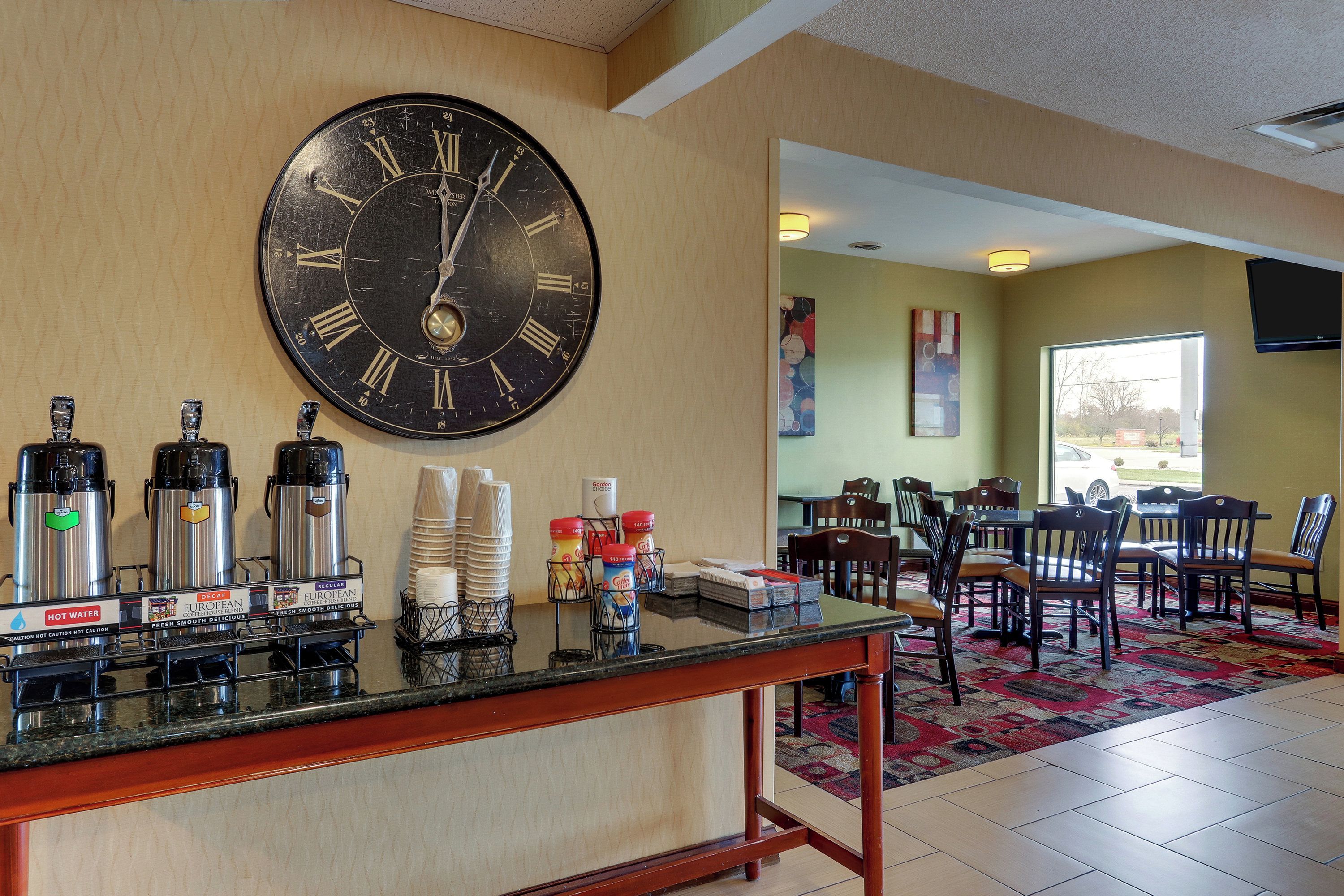 A hotel lobby with a large wall clock, coffee machines, and a seating area with tables and chairs.