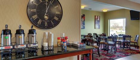 A hotel lobby with a large wall clock, coffee machines, and a seating area with tables and chairs.