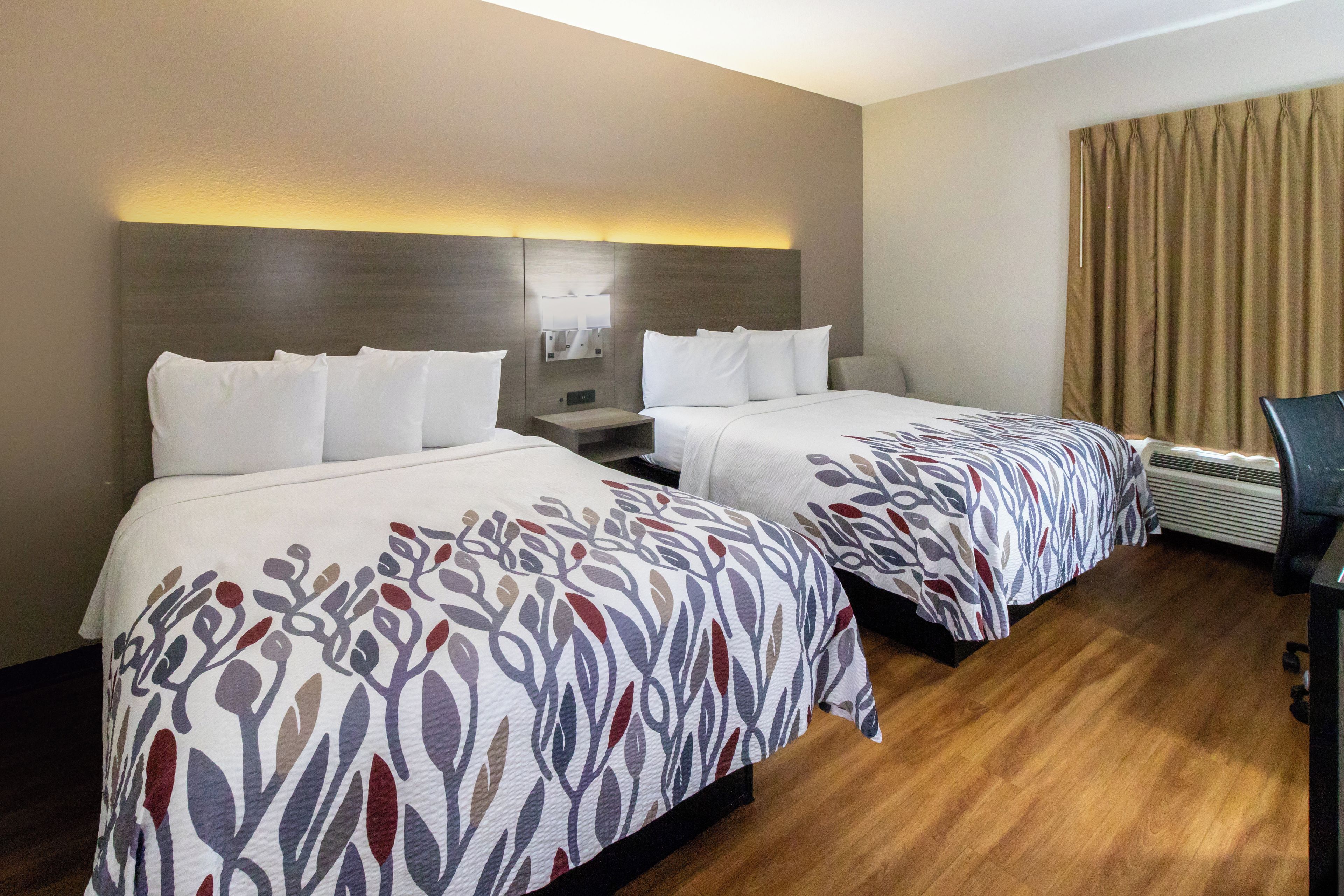 A hotel room with two beds, a nightstand, a wall-mounted lamp, an air conditioning unit, and a curtain.