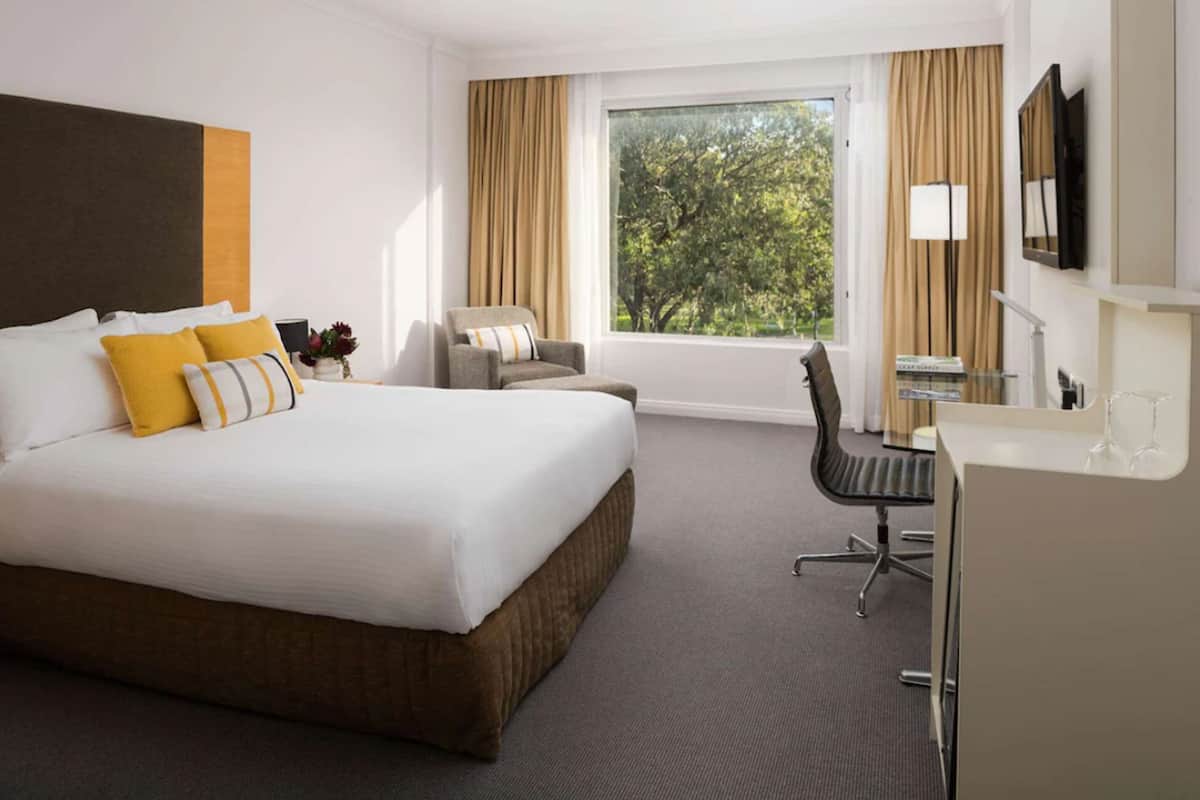 Ett hotellrum med en stor säng, ett skrivbord, en stol och ett fönster med utsikt över träd.