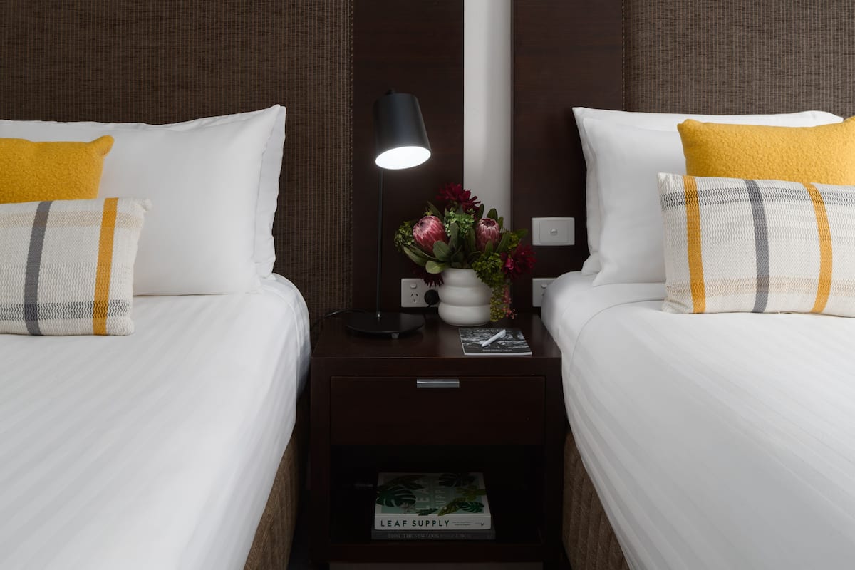 Ett hotellrum med två sängar, båda med vita sängkläder och gula kuddar. Mellan sängarna står ett nattduksbord med en lampa, en vas med blommor och en bok med titeln "Leaf Supply".