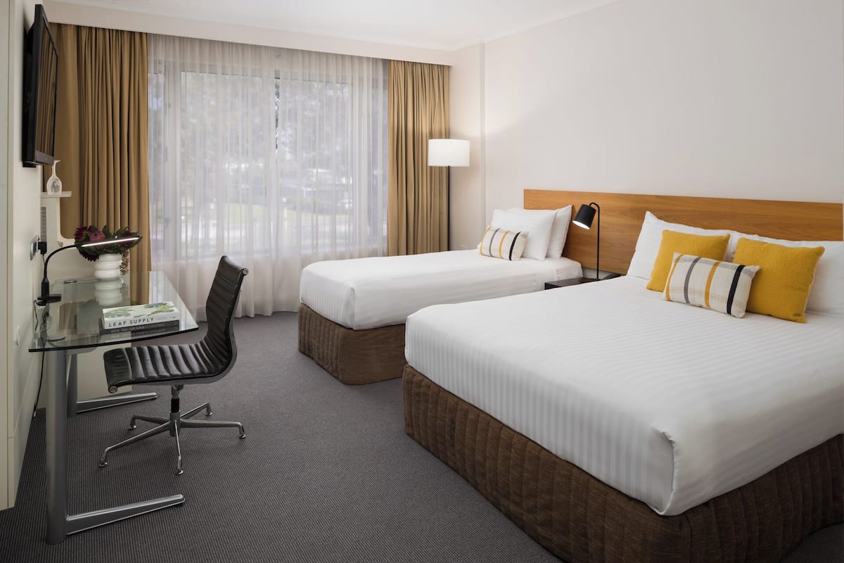 Ett hotellrum med två sängar, ett skrivbord med en stol, en lampa och utsikt genom tunna gardiner.