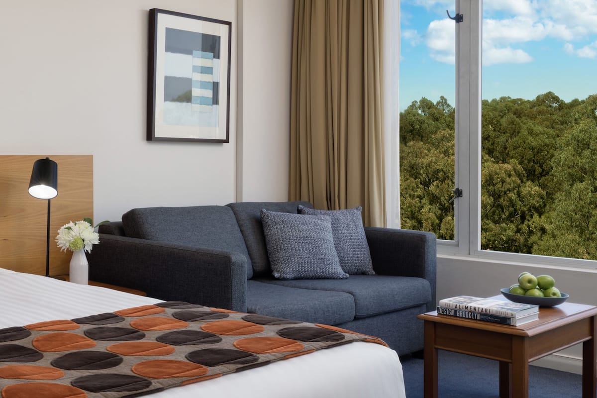 Ett hotellrum med en säng, en grå soffa, ett litet bord med en skål med äpplen och ett fönster med utsikt över träd.