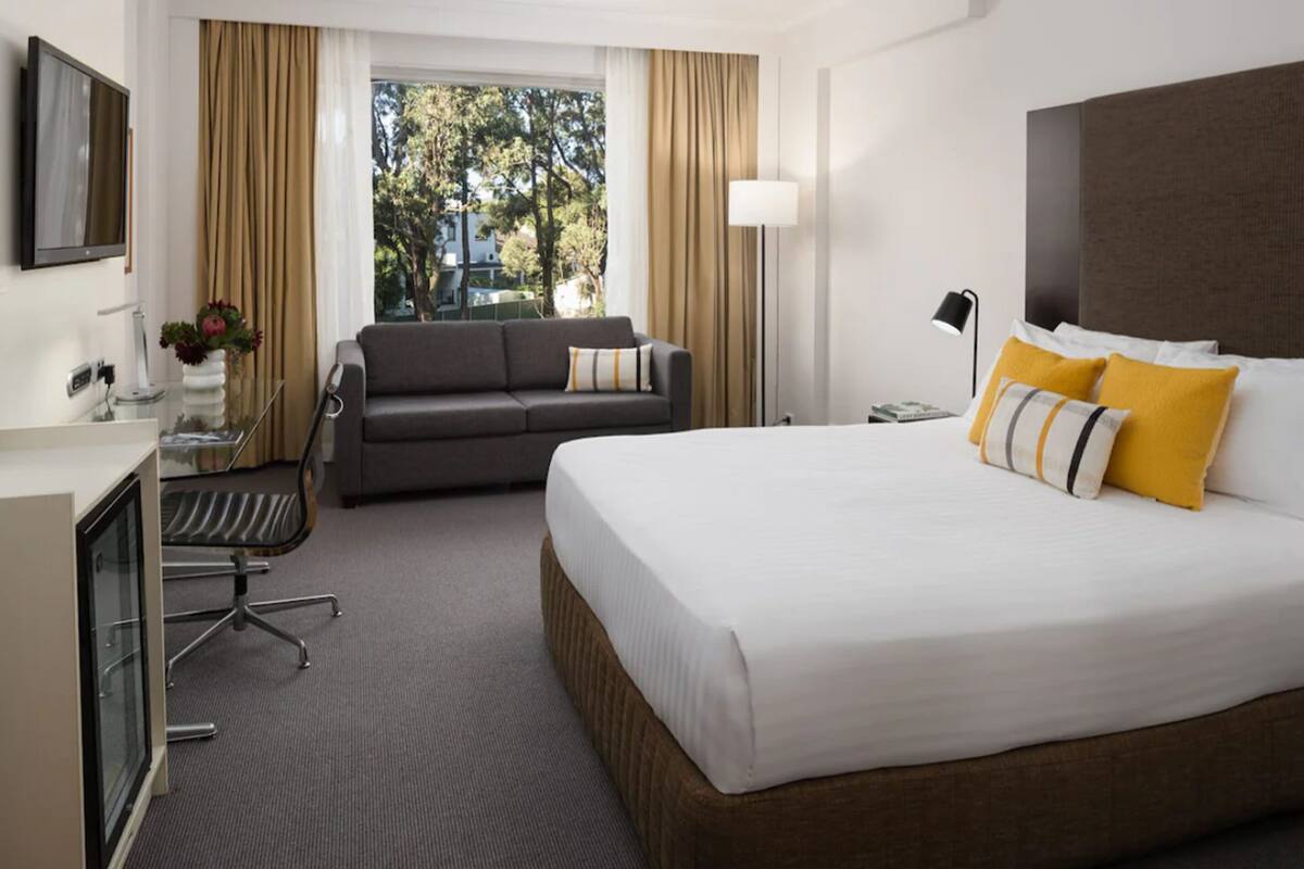 Ett hotellrum med en säng, en soffa, ett skrivbord med en stol, en tv och ett fönster med gardiner.