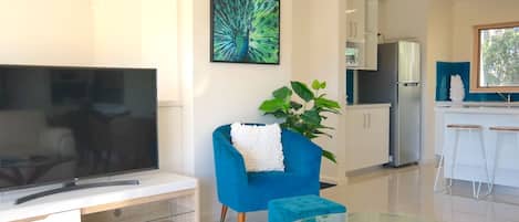 A modern living room with a flat-screen TV, a blue armchair, a glass coffee table, and a kitchen area in the background.