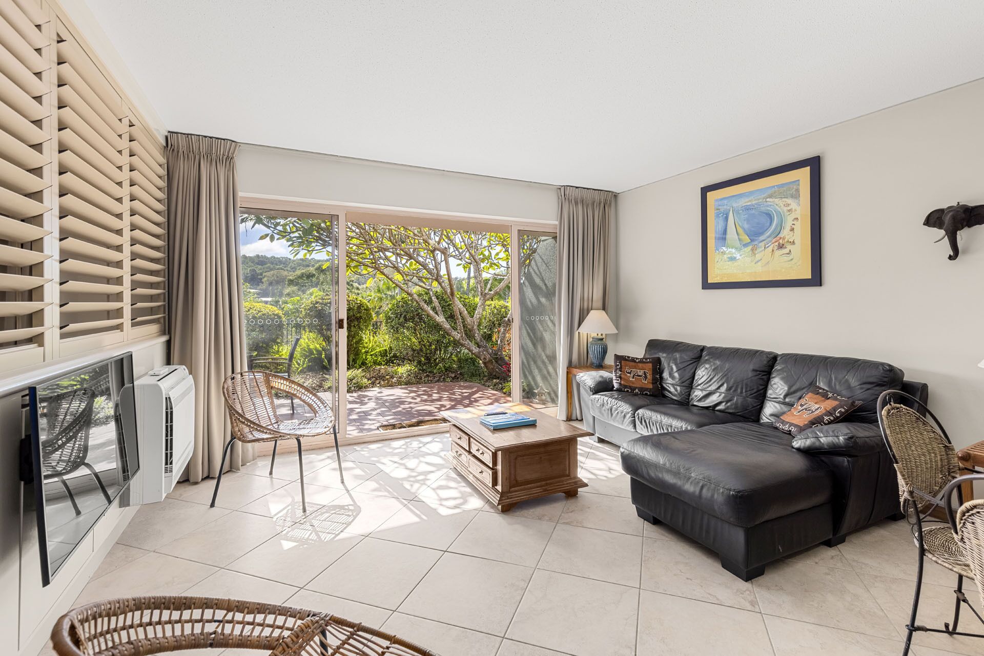 A living room with a sofa, a coffee table, a TV, and a view of a garden through a sliding glass door.