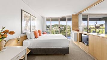 A modern hotel room with a large bed, a desk, and a view of the city through large windows.