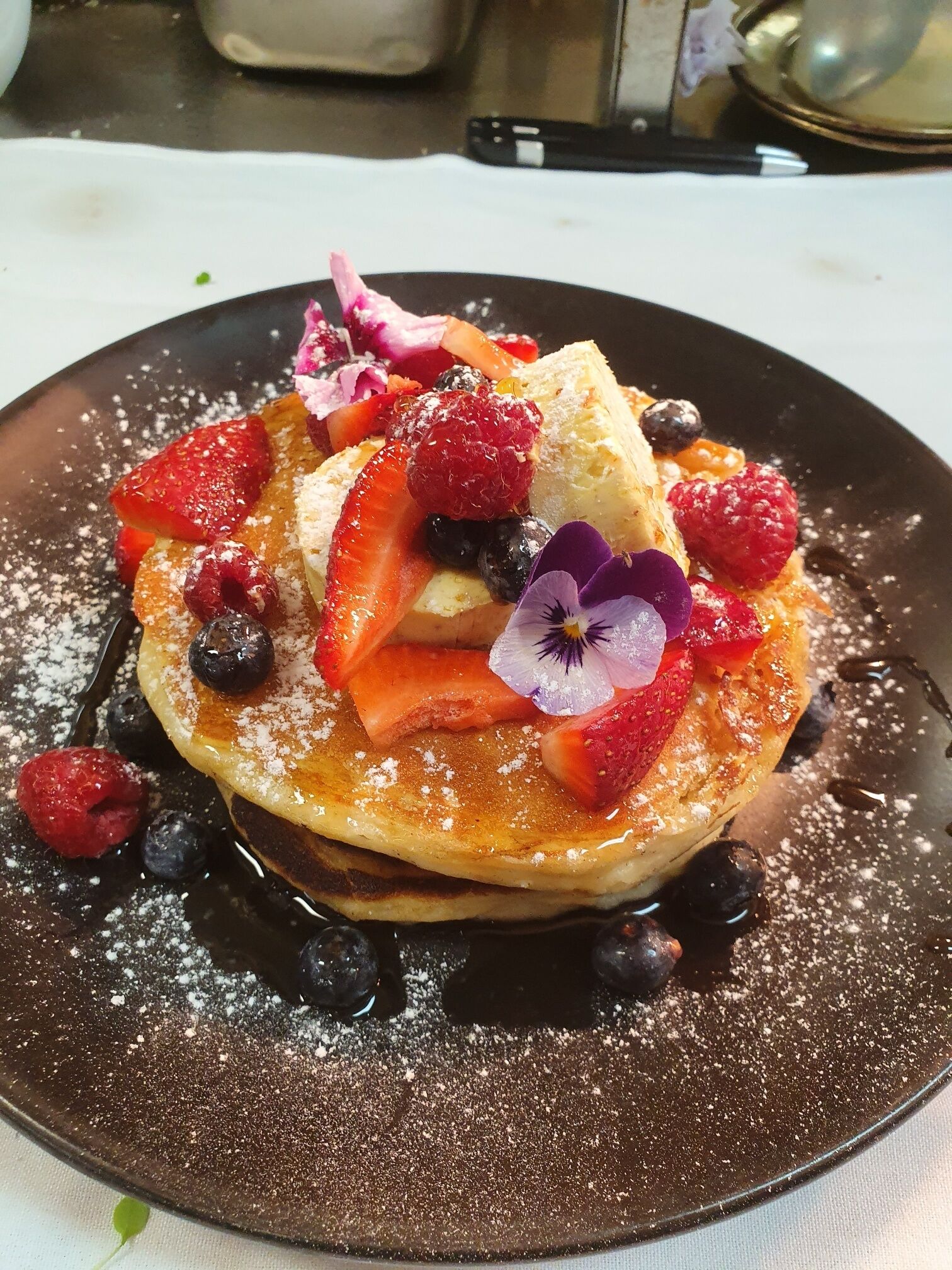 A stack of pancakes topped with berries, powdered sugar, and a flower.