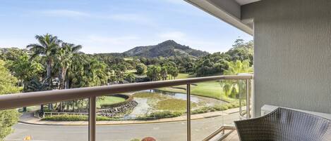 A balcony with a view of a golf course, a mountain, and lush greenery.