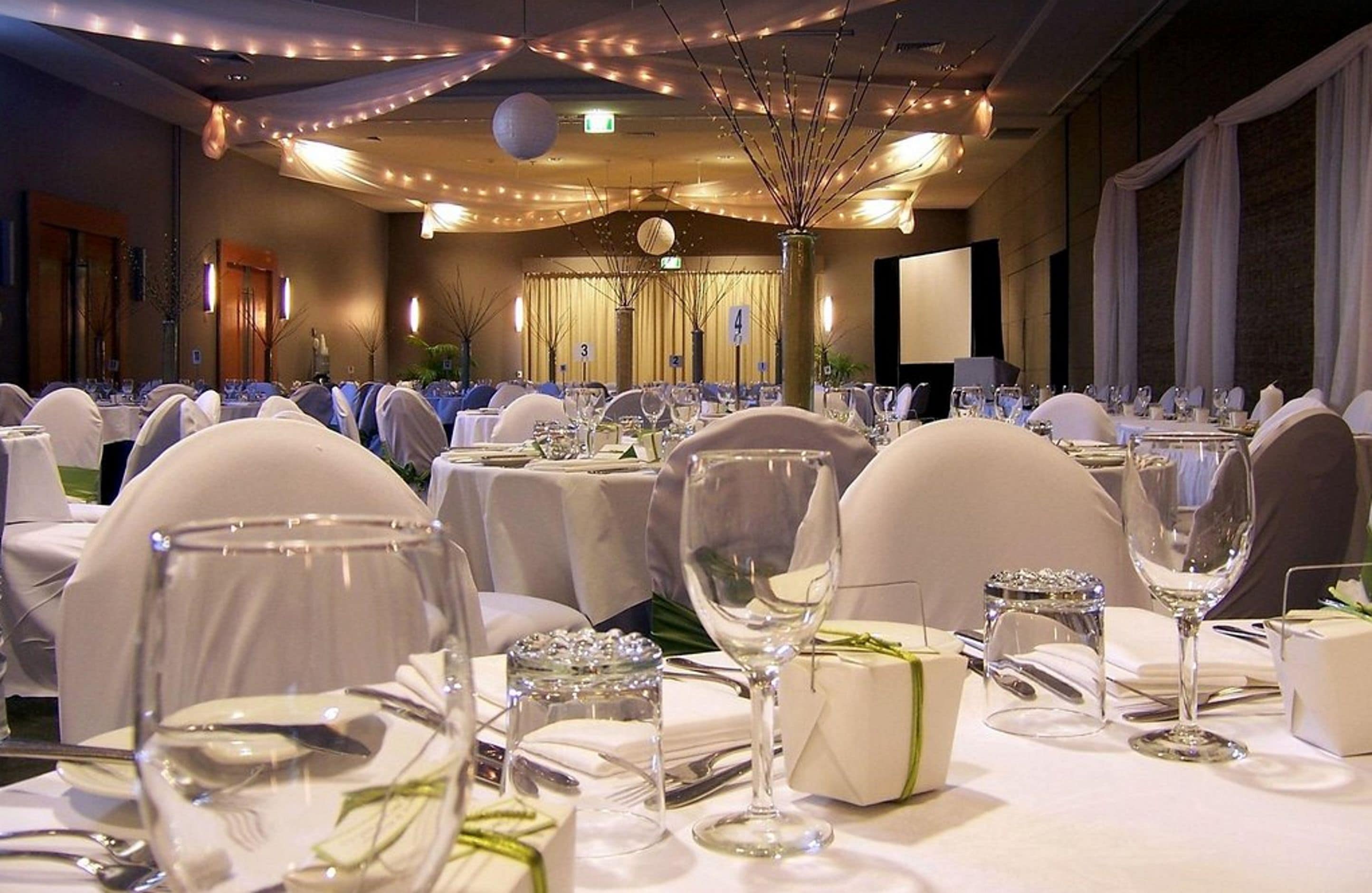 Une salle de banquet avec des tables rondes dressées pour un événement formel, comprenant des nappes blanches, de la verrerie et des chaises.