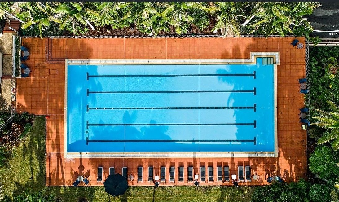 An aerial view of a rectangular swimming pool with multiple lanes, surrounded by a tiled deck and palm trees.