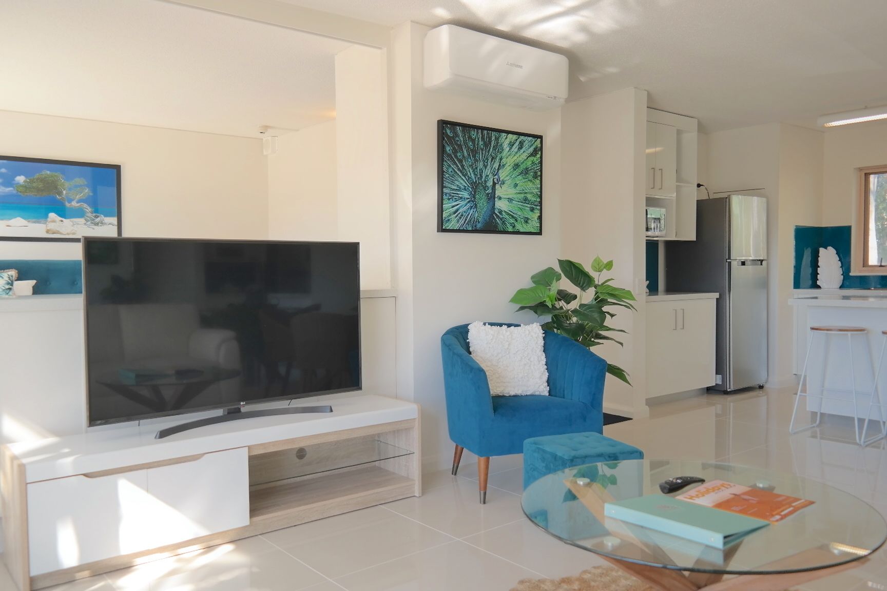A modern living room with a flat-screen TV, a blue armchair, a glass coffee table, and a kitchen area in the background.