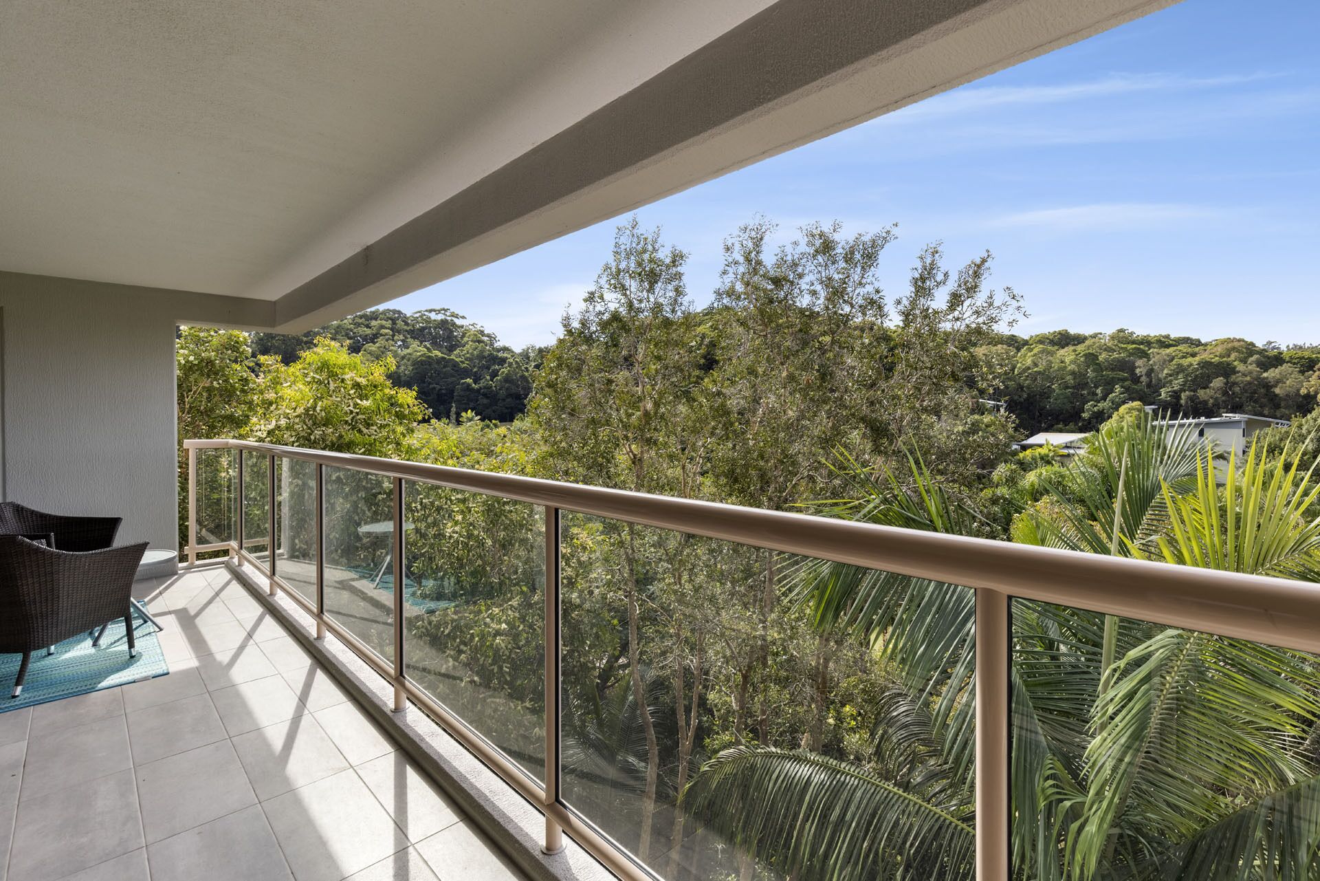 A balcony with a glass railing, a wicker chair, and a view of a lush green landscape with trees and a hill in the background.