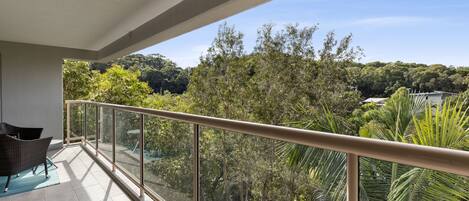 Un balcon doté d’une balustrade en verre, d’un fauteuil en osier et offrant une vue sur un paysage verdoyant, avec des arbres et une colline en arrière-plan.