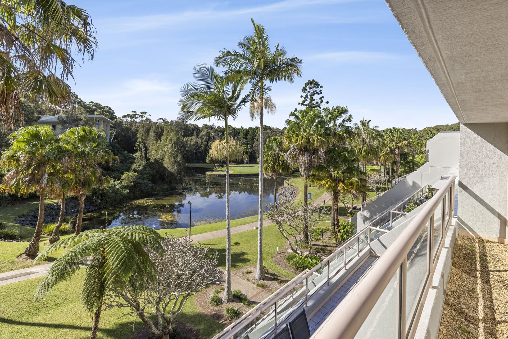 A view from a balcony overlooking a landscaped area with a pond, palm trees, and a golf course in the background.