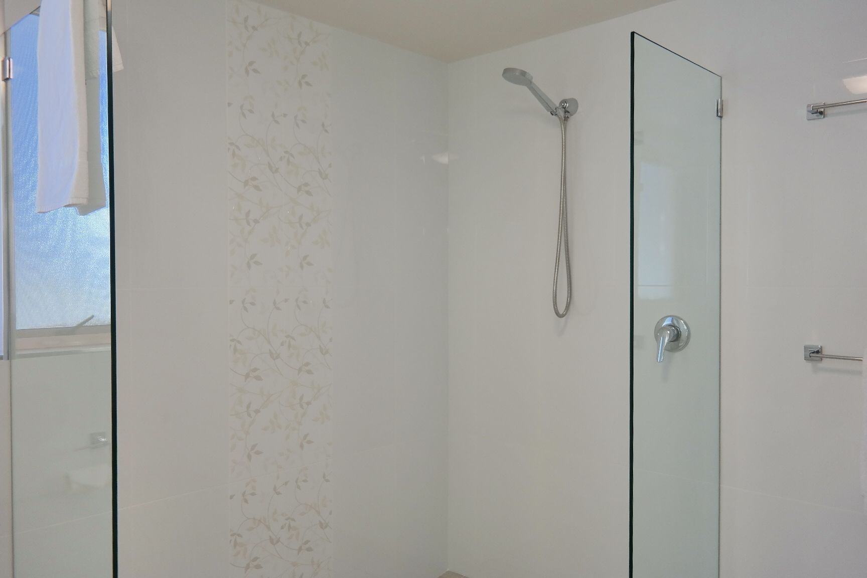 Ducha envuelta en vidrio con cabezal de ducha de mano, puerta de cristal y un perchero visible a la izquierda.