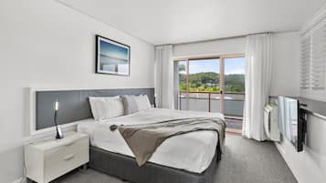 Habitación de hotel moderna con una cama grande, una mesita de noche, un televisor y un balcón con vistas a zonas verdes.