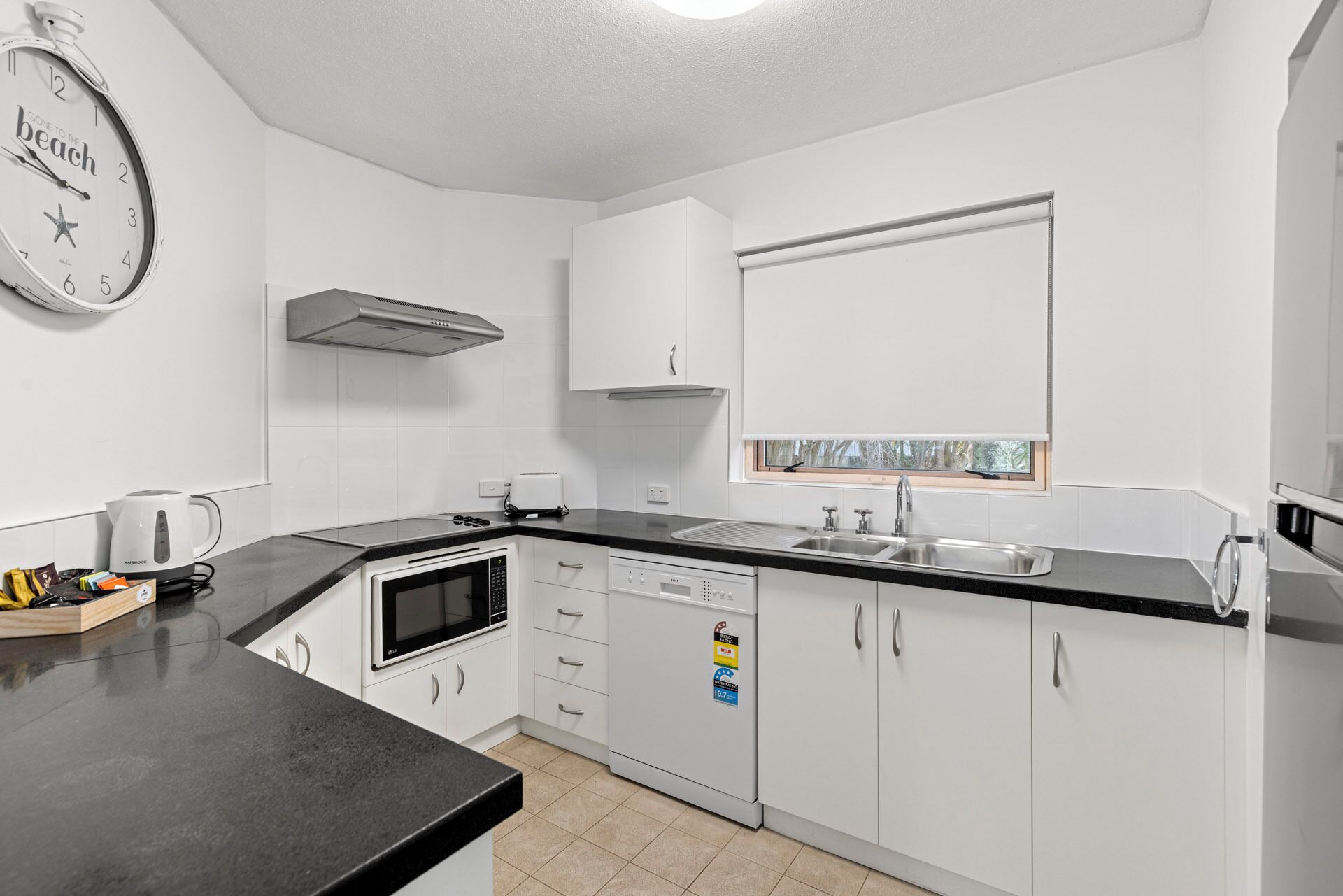 A modern kitchen with white cabinets, a black countertop, a built-in microwave, a kettle, and a window above the sink.