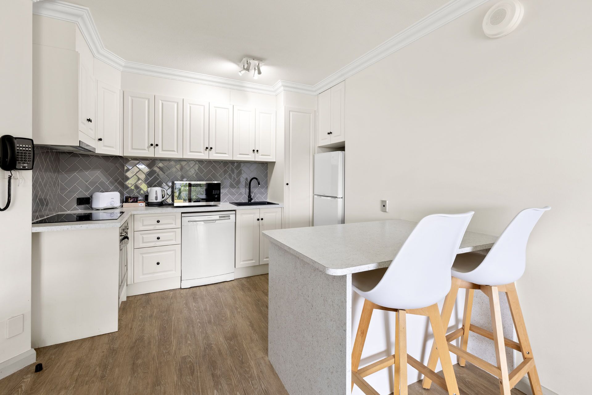 Una cocina moderna con armarios blancos, microondas integrado, refrigerador y cafetera. Hay dos taburetes blancos en la isla.