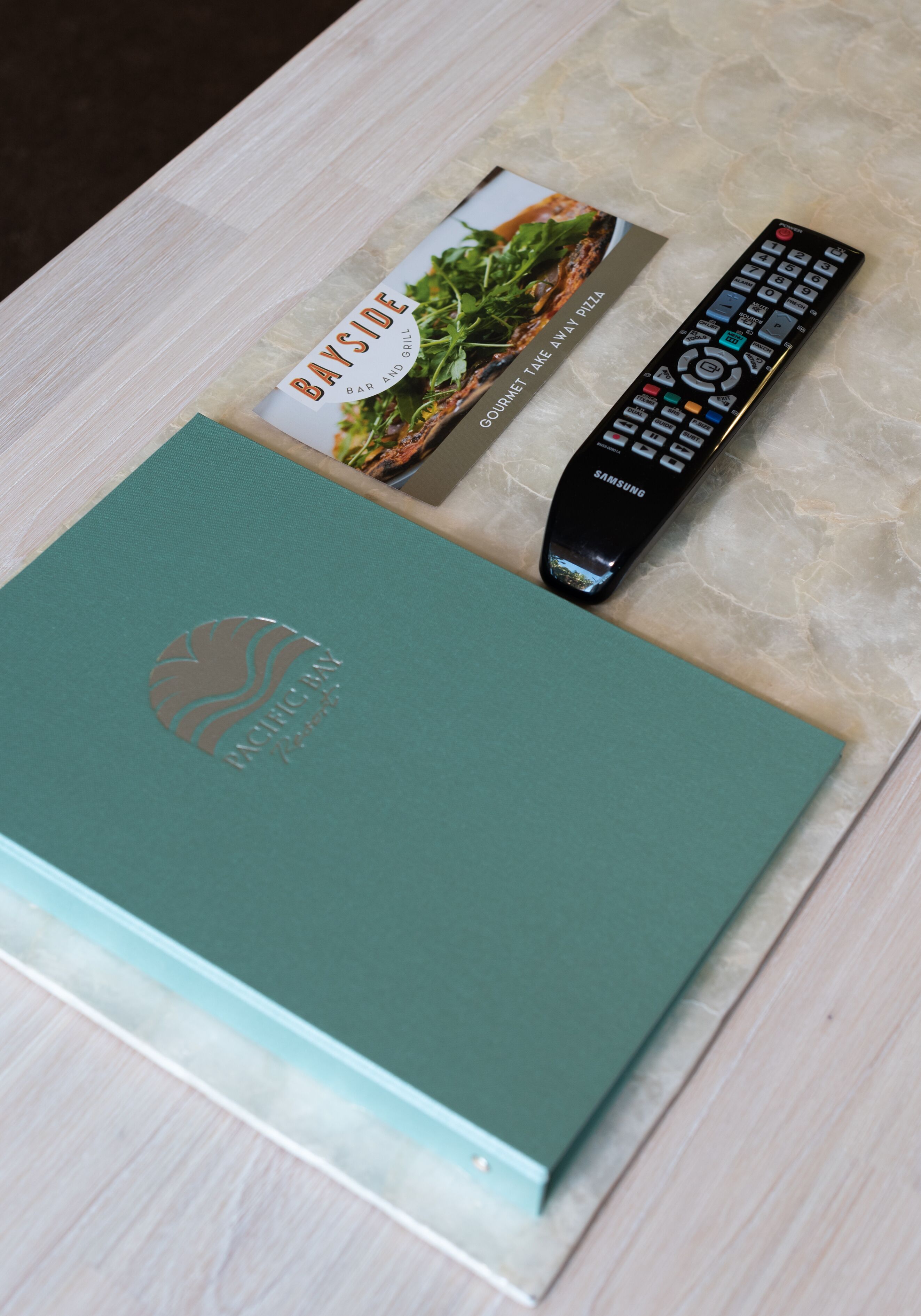 A teal-colored book with a logo, a remote control, and a menu card on a wooden surface.