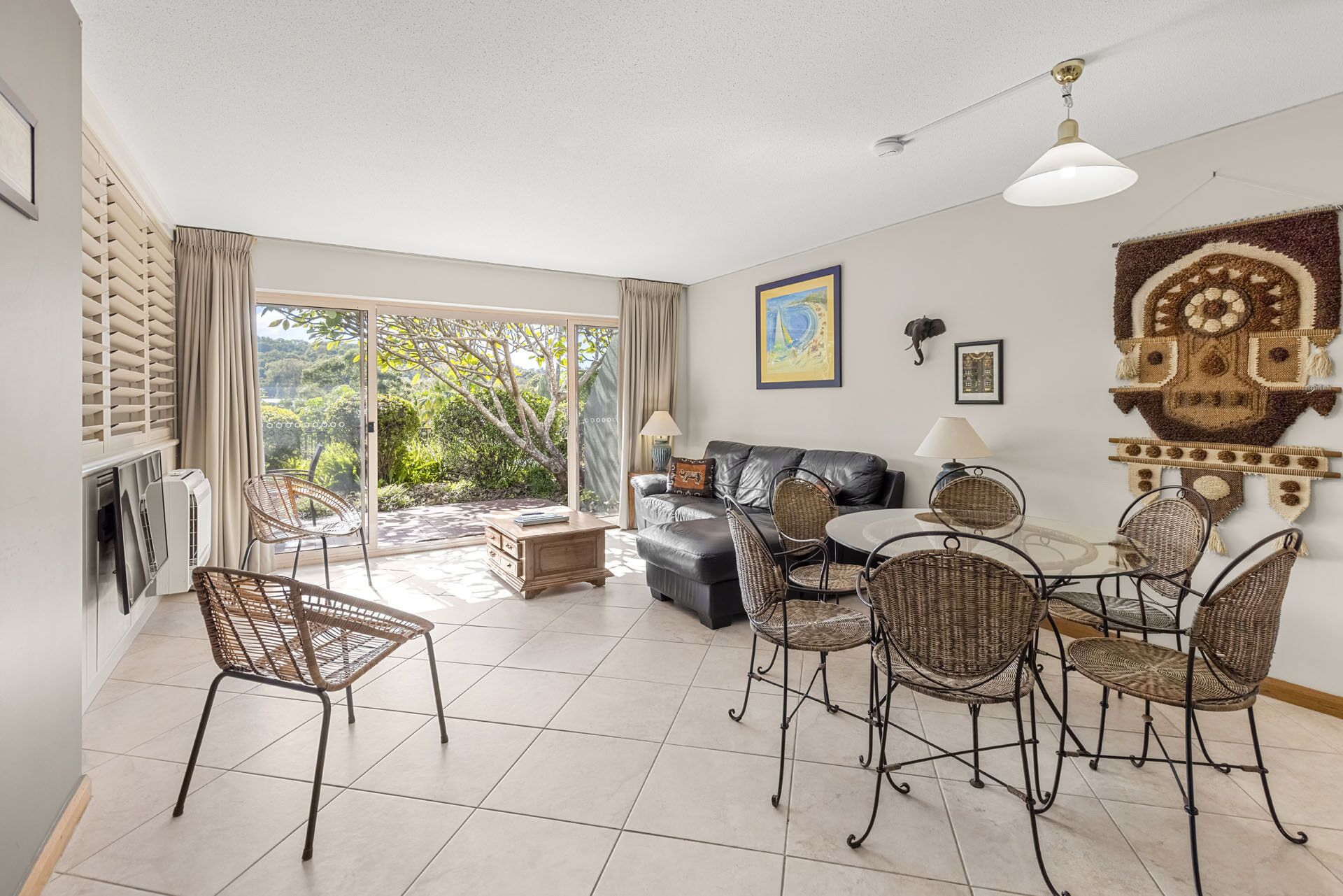 A spacious living room with a sofa, dining table, and chairs, a large window with a view of greenery, and a ceiling fan.