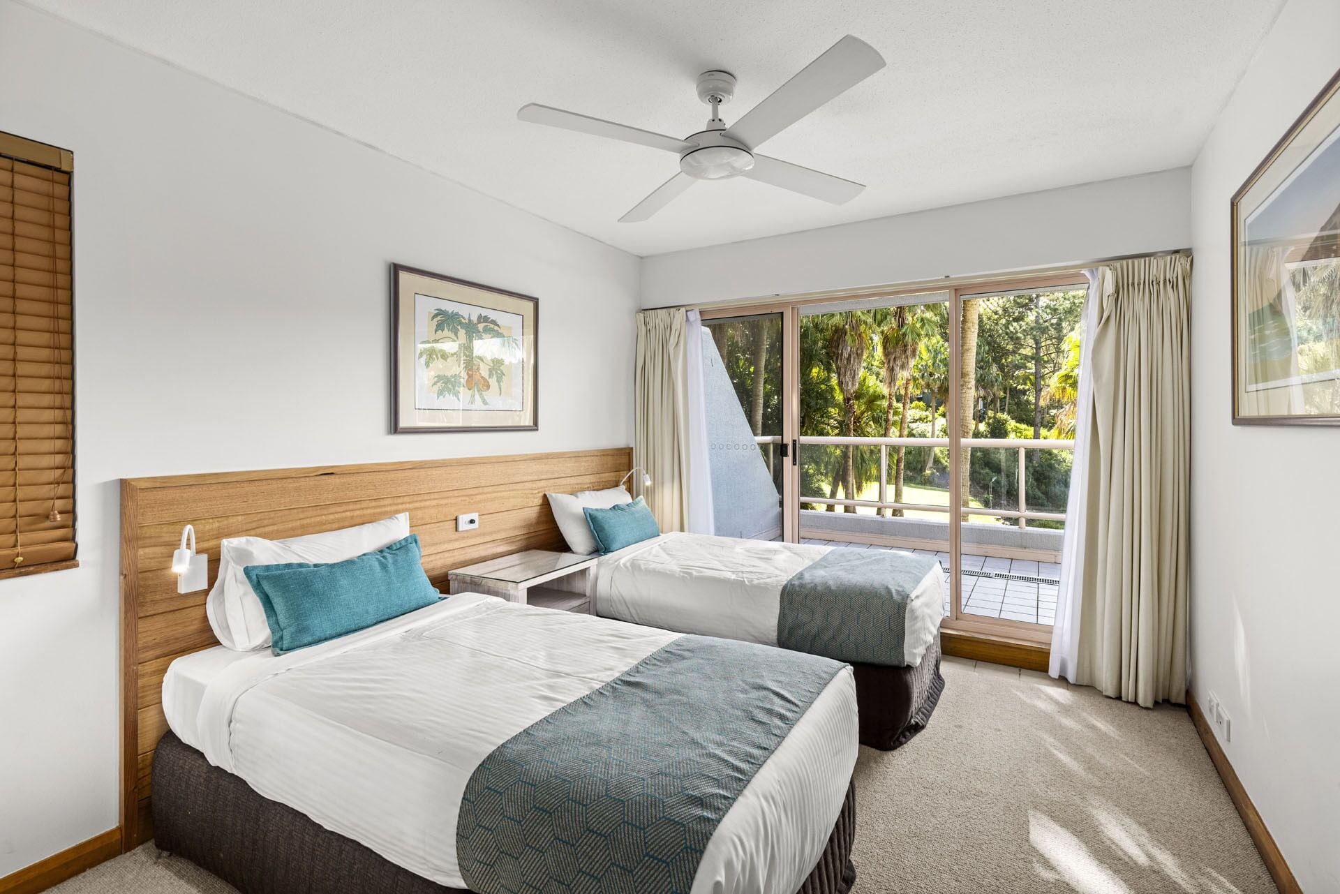 A hotel room with two beds, a ceiling fan, a large window with curtains, and a framed picture on the wall.