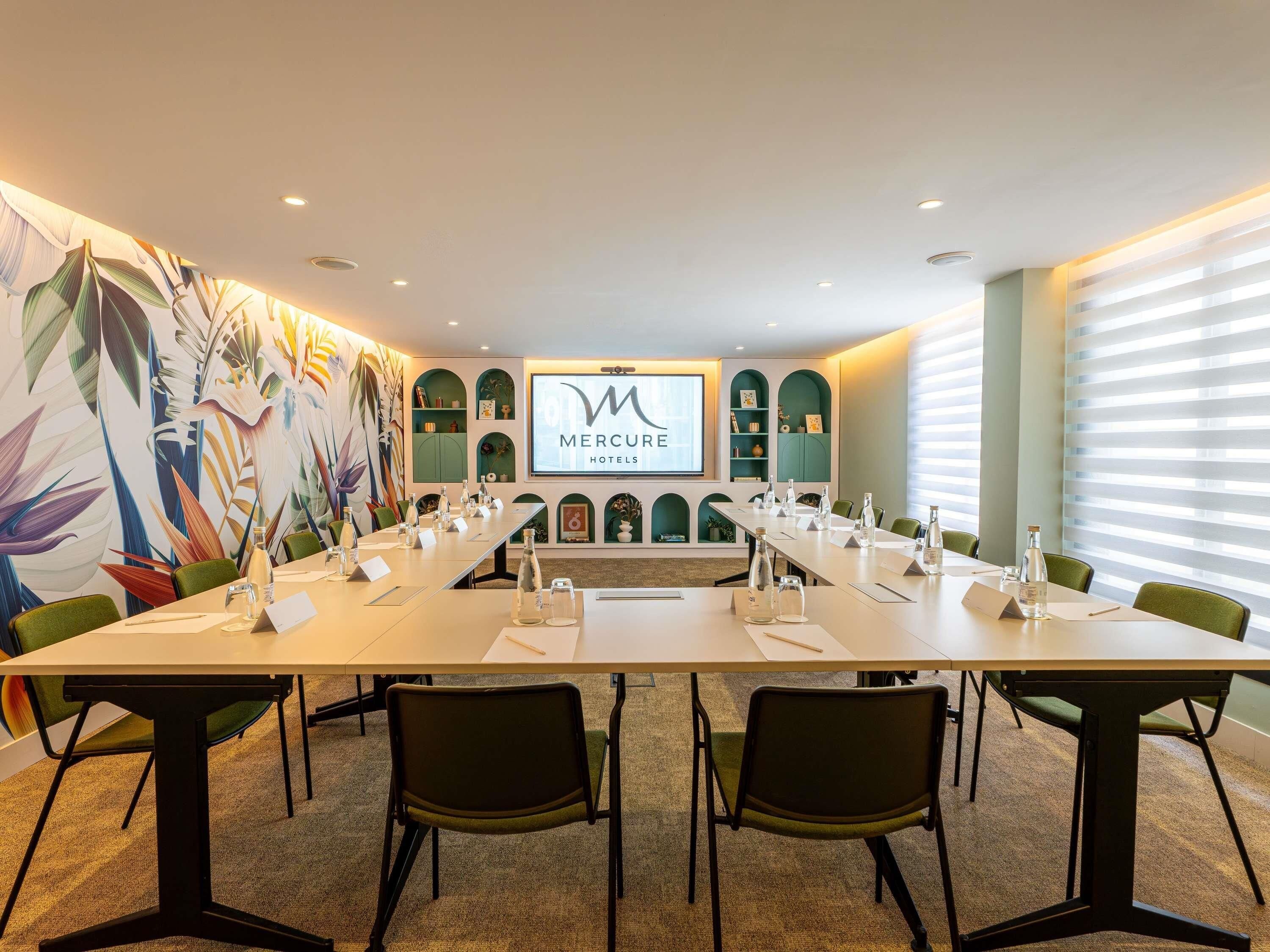 Une salle de conférence équipée d’une longue table, de chaises, d’un écran de projection et d’une fresque représentant des feuilles tropicales.