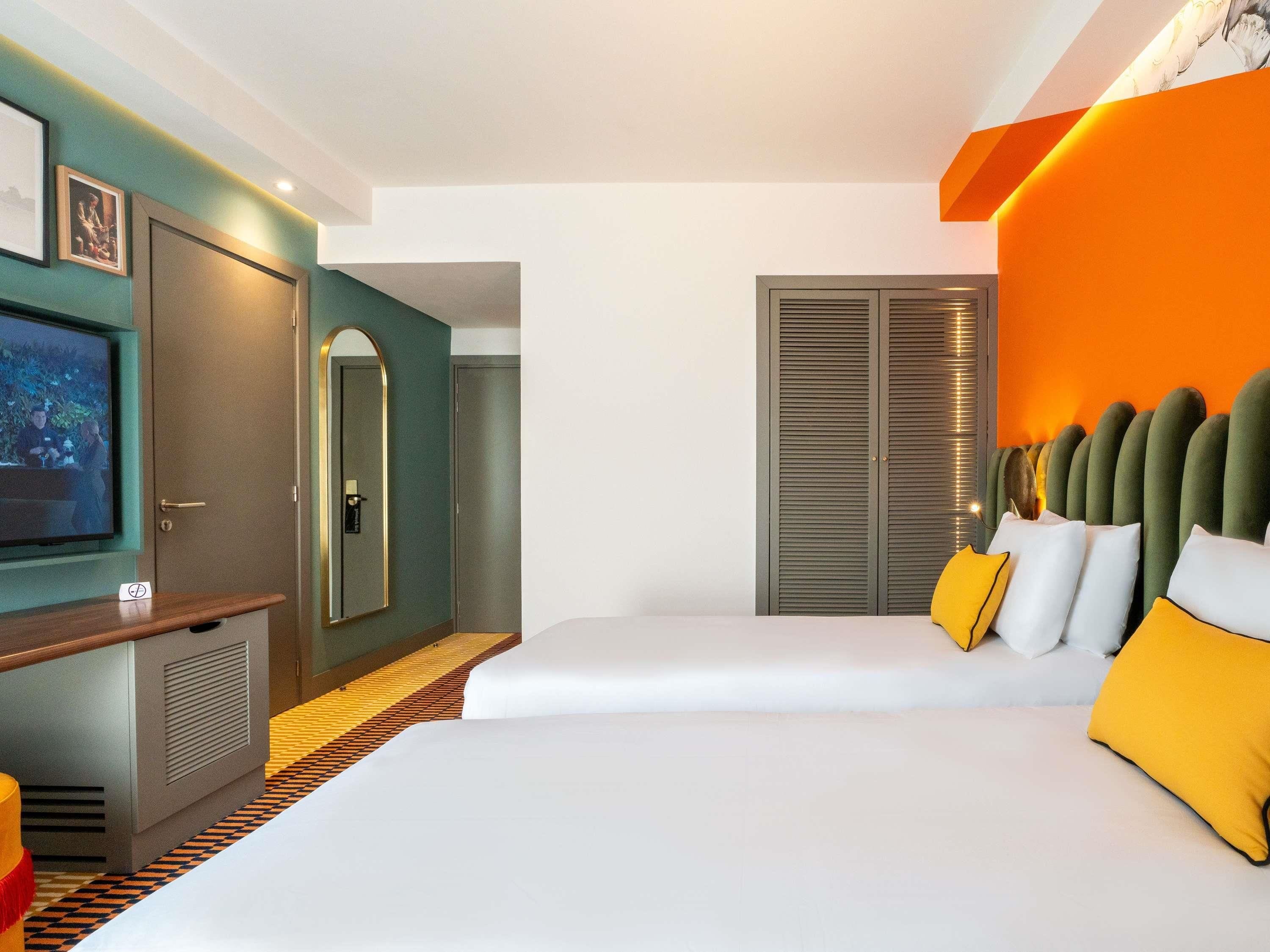 A hotel room with a television, a desk, a bed with yellow pillows, and a wardrobe.