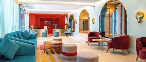A modern hotel lobby with a sofa, ottomans, and chairs, a reception desk, and a red accent wall.