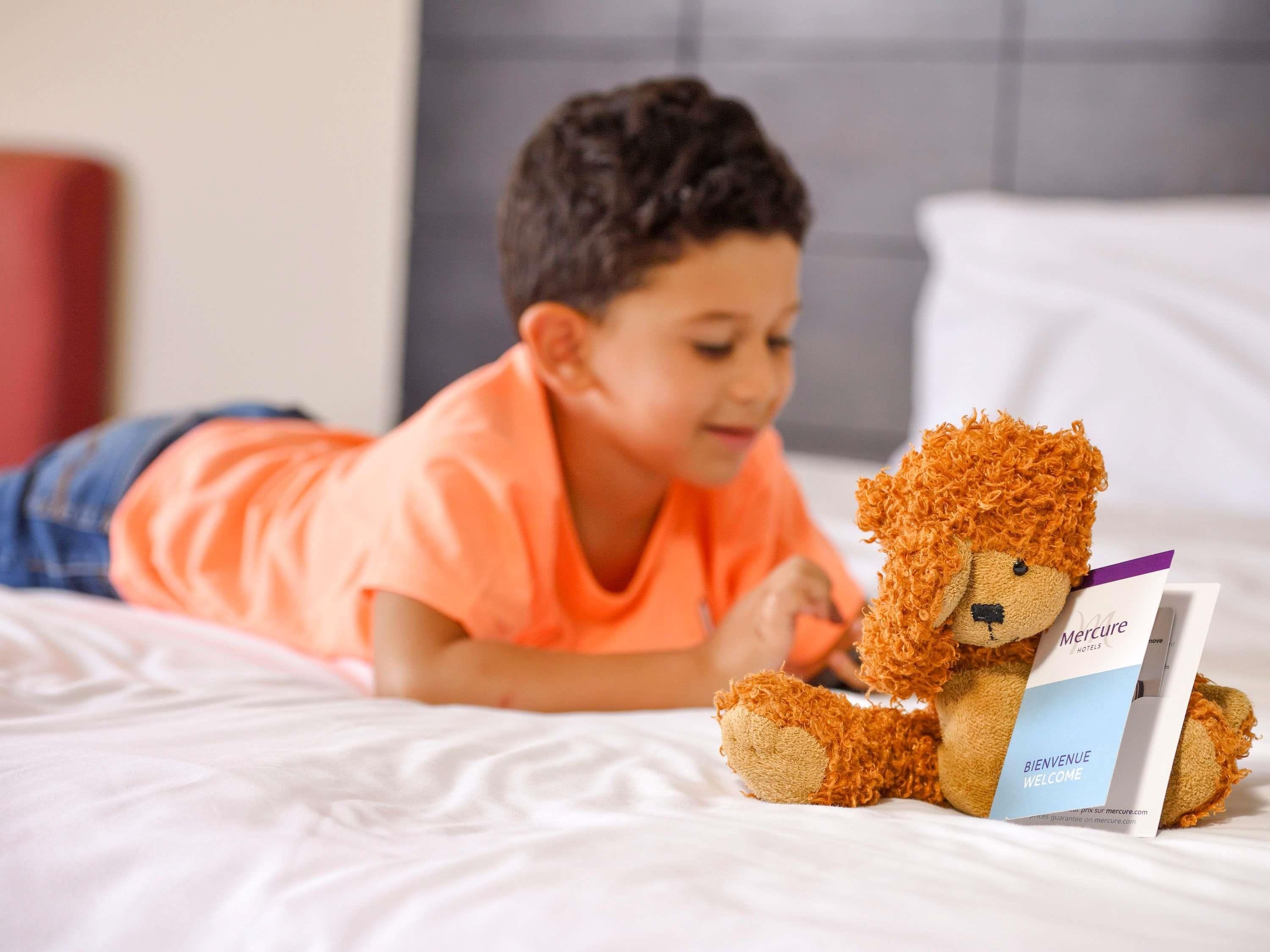 Un niño acostado en una cama con un peluche y una tarjeta de bienvenida del hotel.