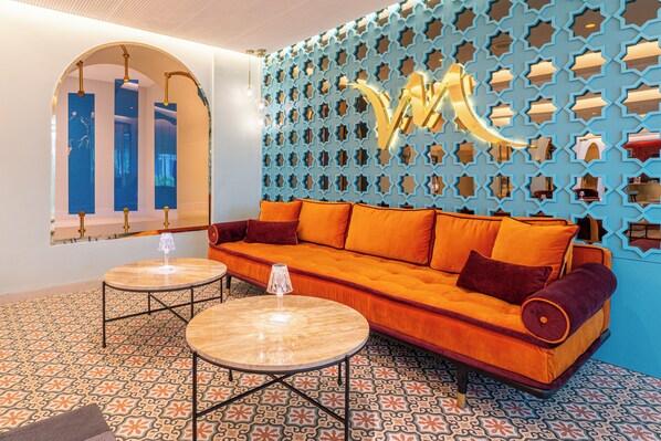 Un intérieur moderne avec un canapé, deux tables rondes, des carreaux de sol à motifs et un mur décoratif orné de formes géométriques.
