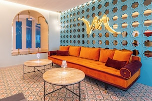 Ein modernes Interieur mit einer Couch, zwei runden Tischen, fußbodenhohen, gemusterten Fliesen und einer Wand mit geometrischen Formen.