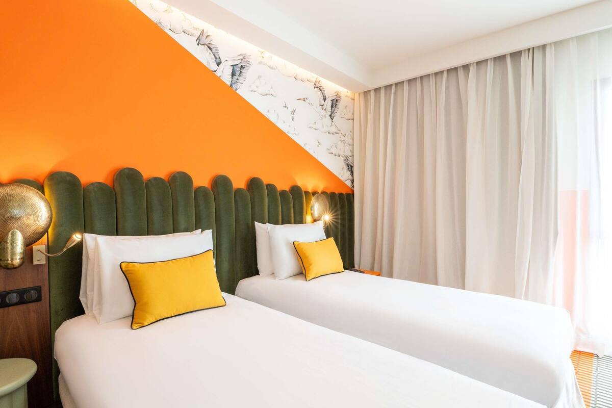 A hotel room with two beds, a green headboard, and a large window with curtains.