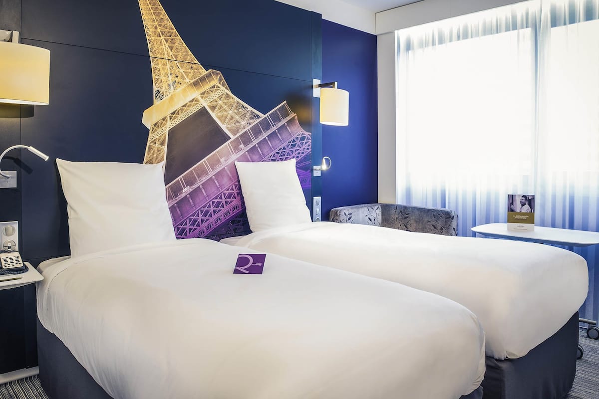 A hotel room with a bed, a nightstand, a lamp, a wall-mounted TV, and a window with curtains.