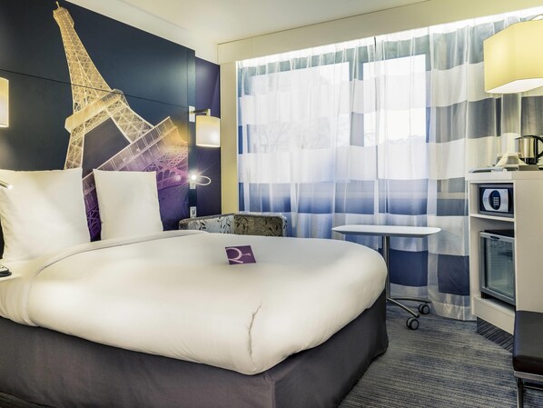 Une chambre d’hôtel comprenant un lit, une petite table, une chaise, un bureau avec un ordinateur, un four à micro-ondes et une grande fenêtre avec des rideaux.