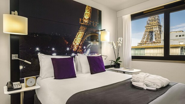 A hotel room with a bed, a nightstand, a lamp, a window with a view of the Eiffel Tower, and a small potted plant. - Mercure Paris Centre Tour Eiffel (Paris)