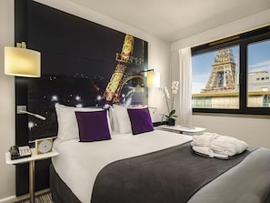 Una habitación de hotel con cama, mesita de noche, lámpara, ventana con vista a la Torre Eiffel y una pequeña planta en maceta.