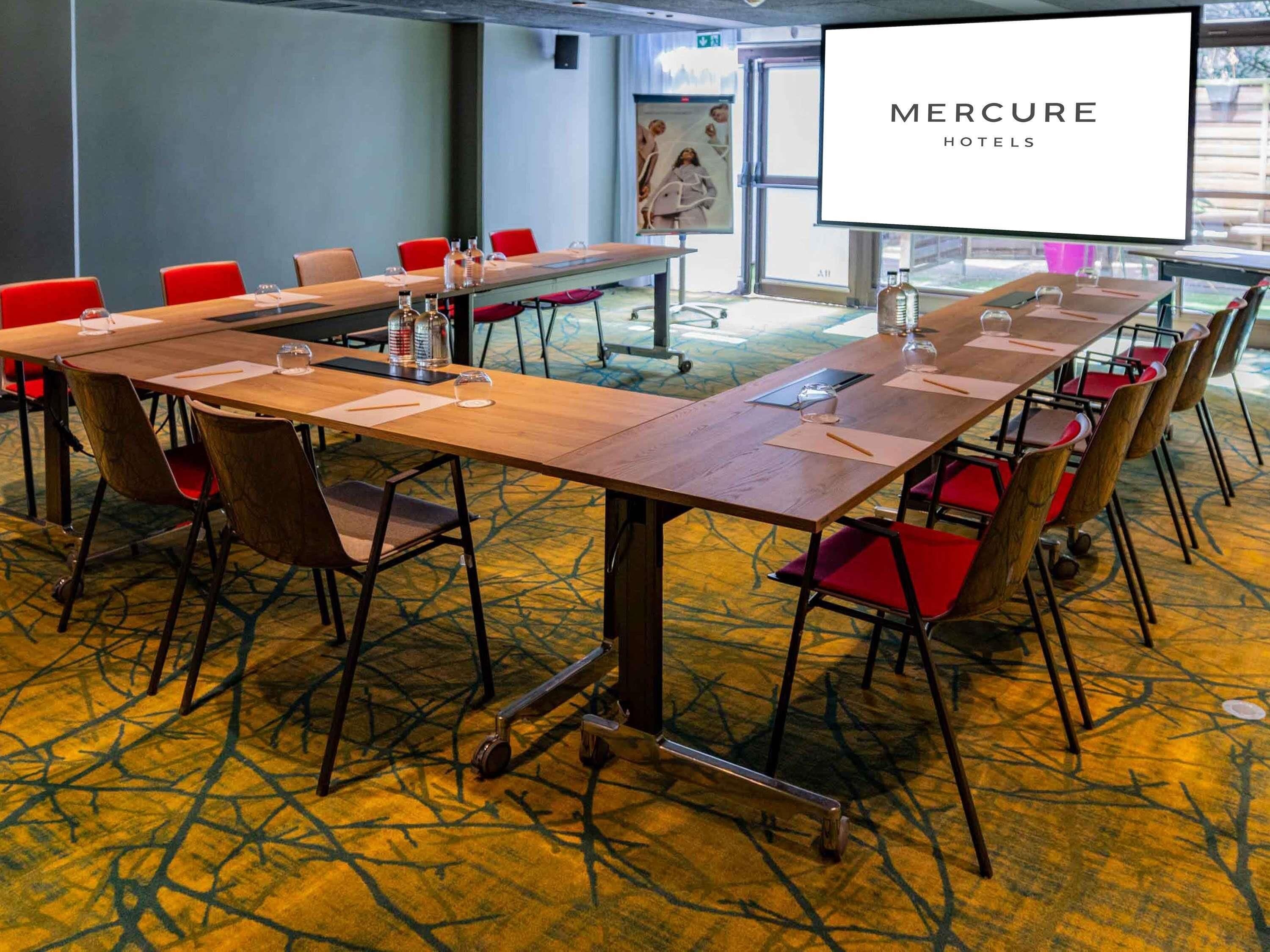 Un salón de conferencias con una larga mesa, sillas rojas y una pantalla proyectora mostrando 'MERCURE HOTELS'.