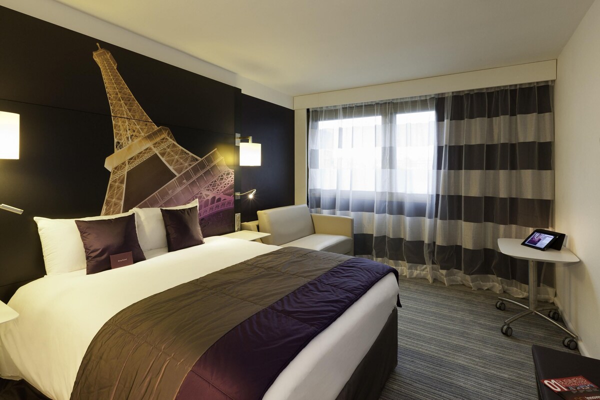 Une chambre d’hôtel comprenant un lit, un bureau, une chaise, une grande fenêtre avec des rideaux et une fresque murale représentant la Tour Eiffel.